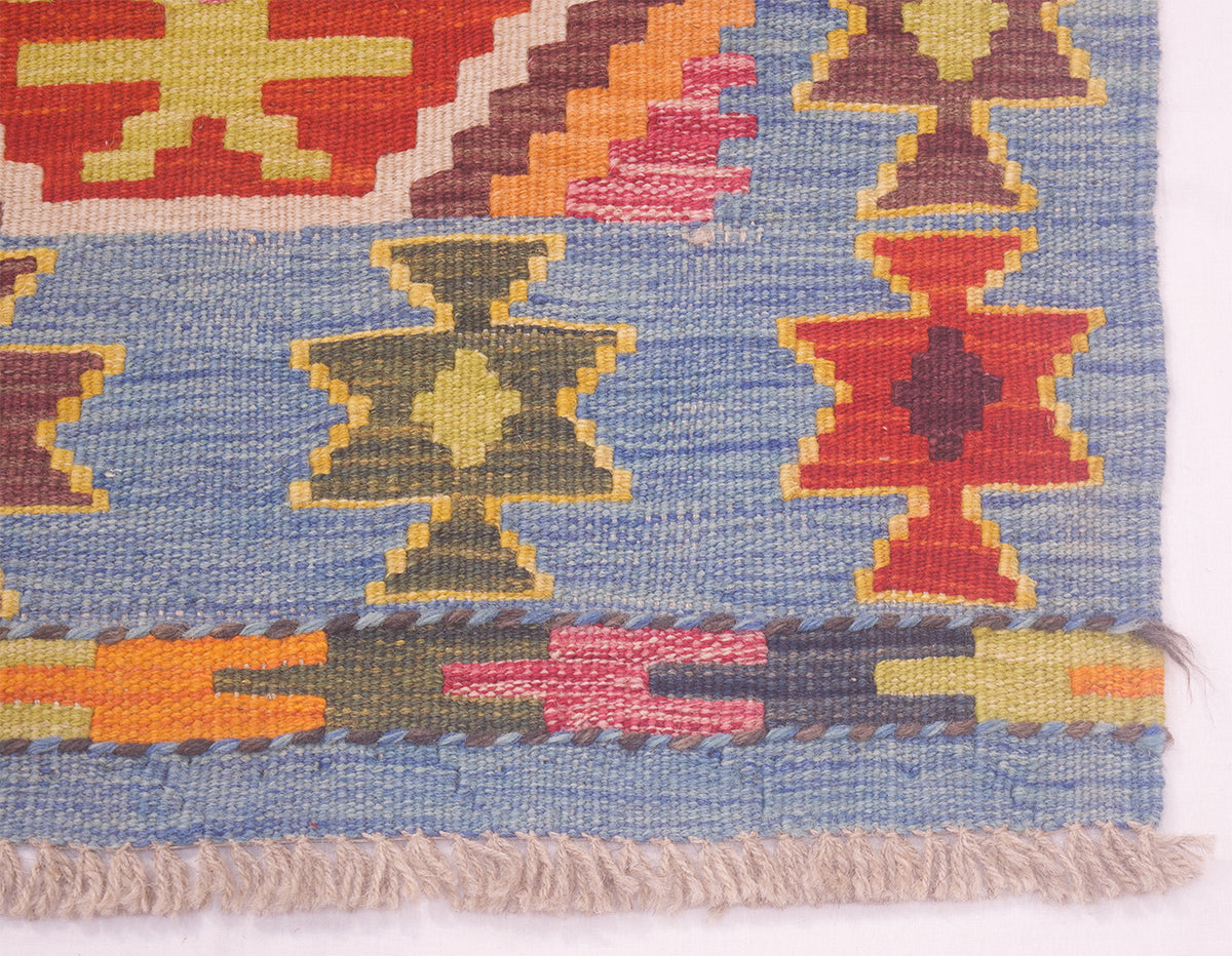 Close-up of a Afghan Kilim mat handwoven with a geometric pattern, featuring a light blue field, red, yellow, olive green, and pink motifs, and a fringed edge.