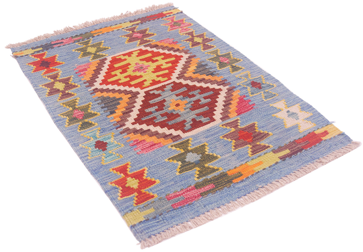 A rectangular woven kilim rug with a geometric pattern, featuring a dusty blue background with red, orange, yellow, and green motifs, finished with fringed edges.
