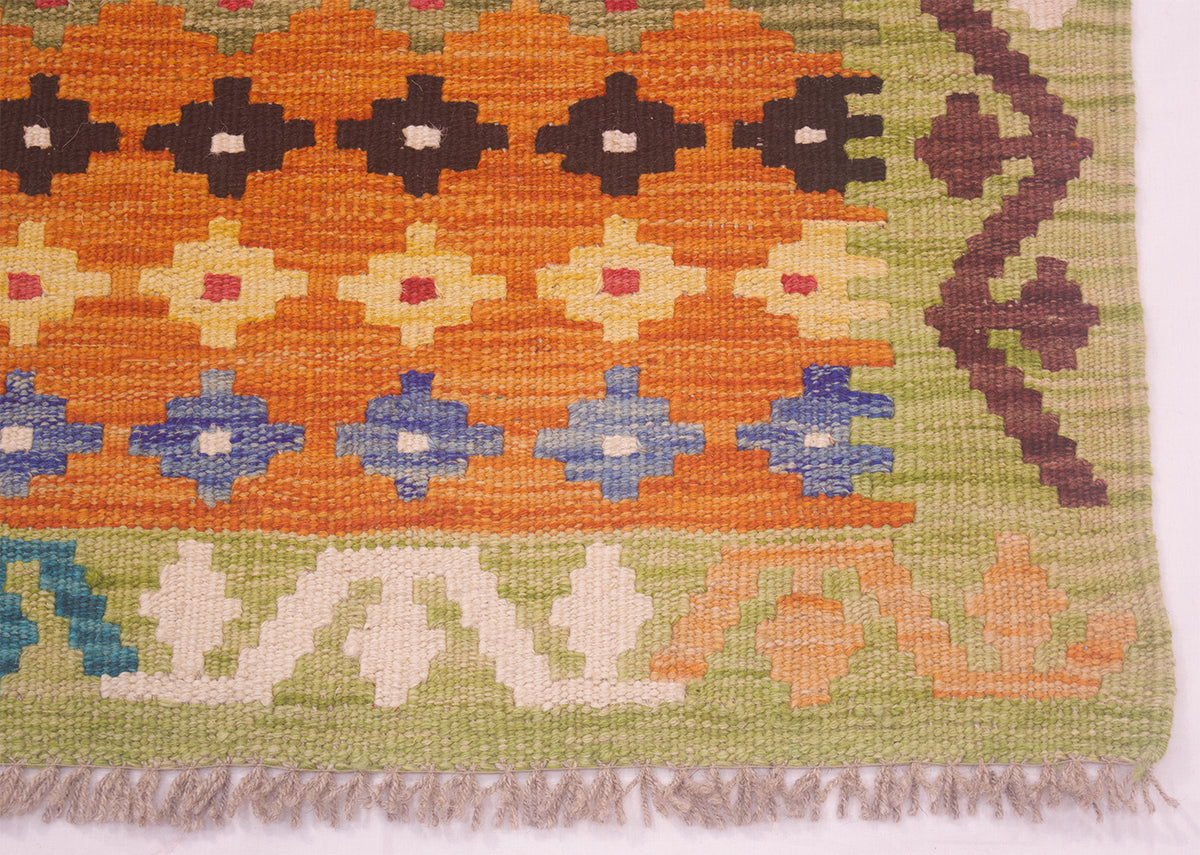 A close-up of a woven kilim rug showing its geometric pattern and fringed edge. The design features a prominent orange center band with dark brown, cream, and blue diamond motifs, bordered by a light olive green section with cream and peach patterns.
