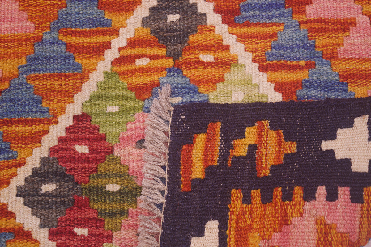 Close-up of a woven geometric Afghan Kilim mat with a multicolour pattern including orange, pink, blue, green, red, and cream, featuring a dark navy corner with cream fringe.