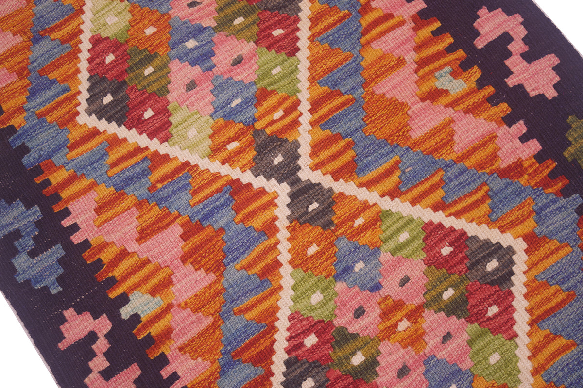 A close-up of a woven kilim rug featuring a vibrant geometric pattern with pink, orange, blue, green, and red shapes outlined in cream, bordered by a dark navy blue edge.