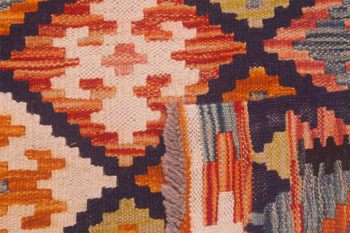 Close-up of Afghan kilim rug showing a geometric pattern in orange, cream, navy with a fringed edge.