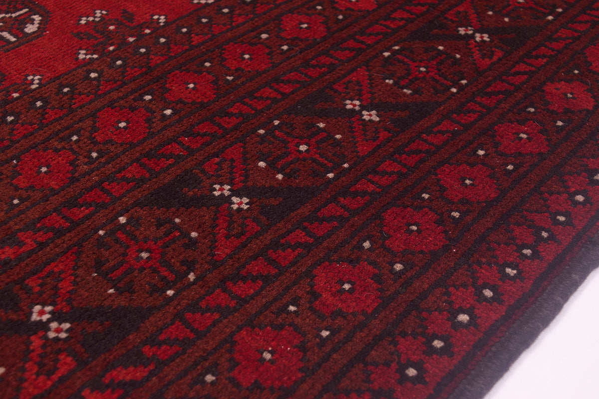 A close-up of a hand-knotted Afghan carpet with deep red and black geometric bands, small white dot accents, and a dark border along one edge.