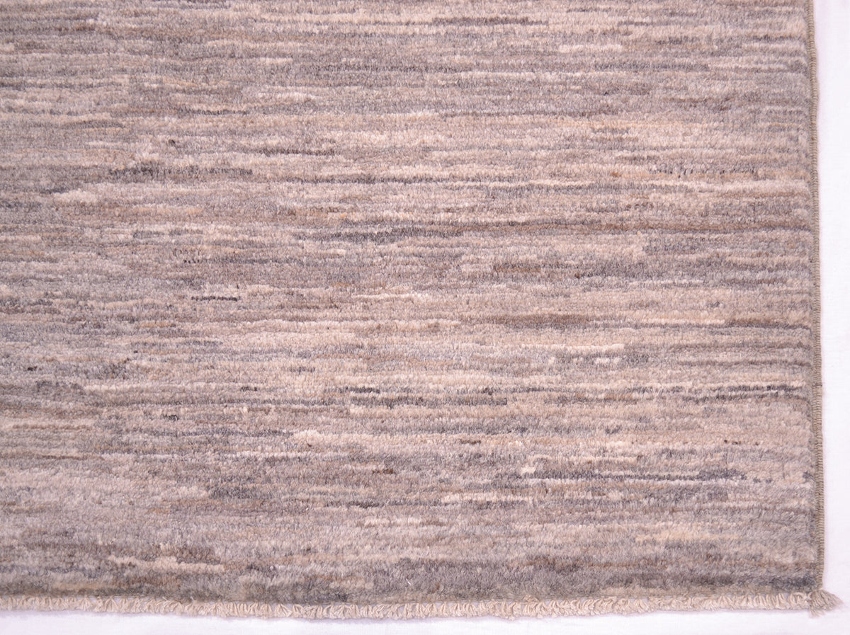 A close-up of a corner of a textured handwoven plain rug with horizontal striations in taupe, beige, and grey tones, showing a frayed edge along the bottom.