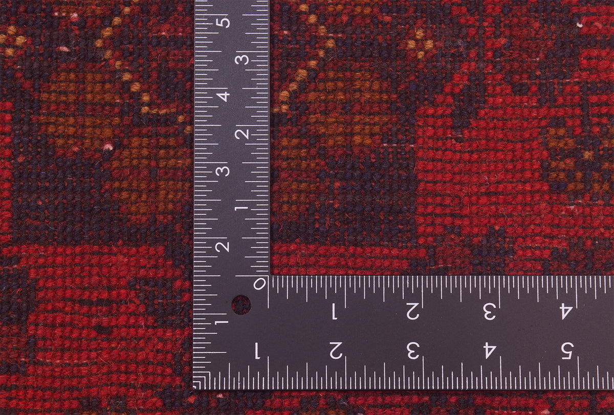 Close-up of back of a patterned runner rug with L-shaped ruler placed over it to show scale, featuring a red, brown, and black woven design.