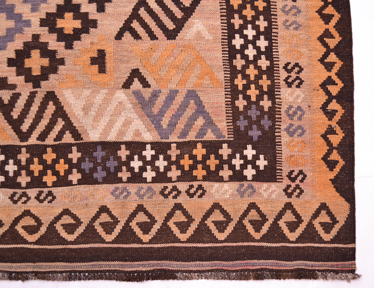 A close-up of a mature colour handwoven large old Kilim rug corner showing dark brown, tan, orange, and muted blue patterned sections with fringe along the bottom edge.