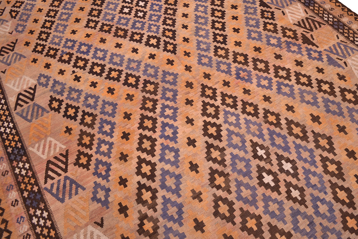 A close-up of a mature colour large old Kilim rug showing a geometric design with repeating diamond and cross motifs in brown, blue, beige, and white on a peach background.