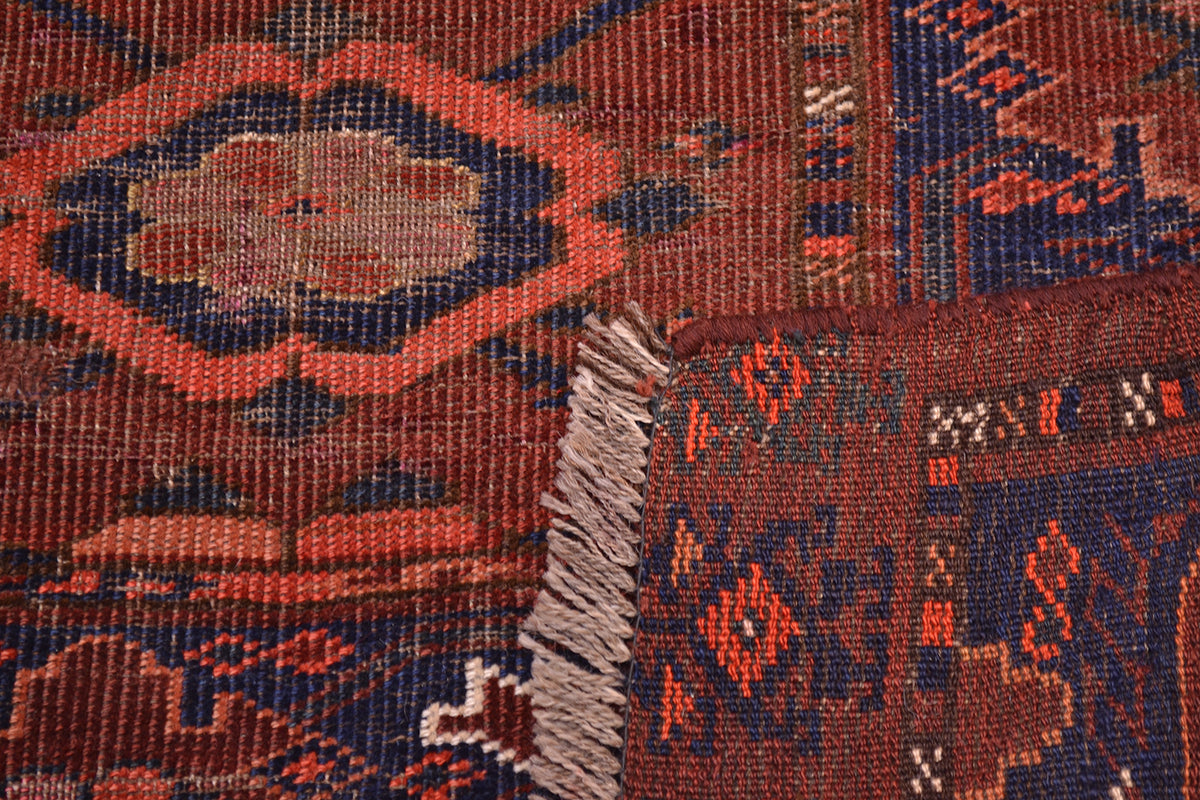 Close-up of the hand-knotted back corner turned up to show the weave of and Antique Ersari Chuval patterned rug showing a rust red field with dark blue geometric motifs and a fringed edge.