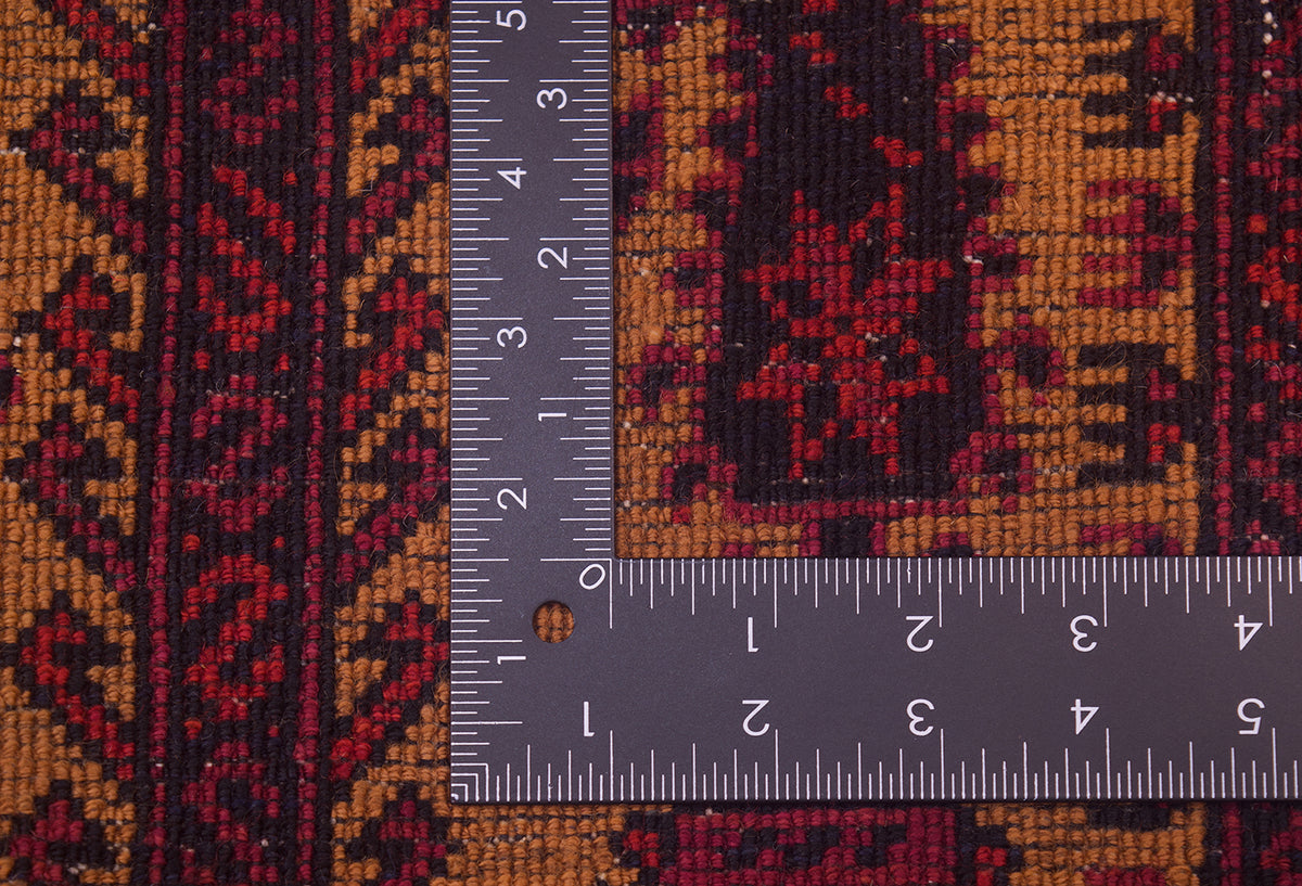 Close-up of the hand-knotted weave of an Antique Belouch prayer rug with a metal L-square ruler placed over it to show scale of the knotting, showing a woven rug in rust orange, red, and dark brown/black tones.
