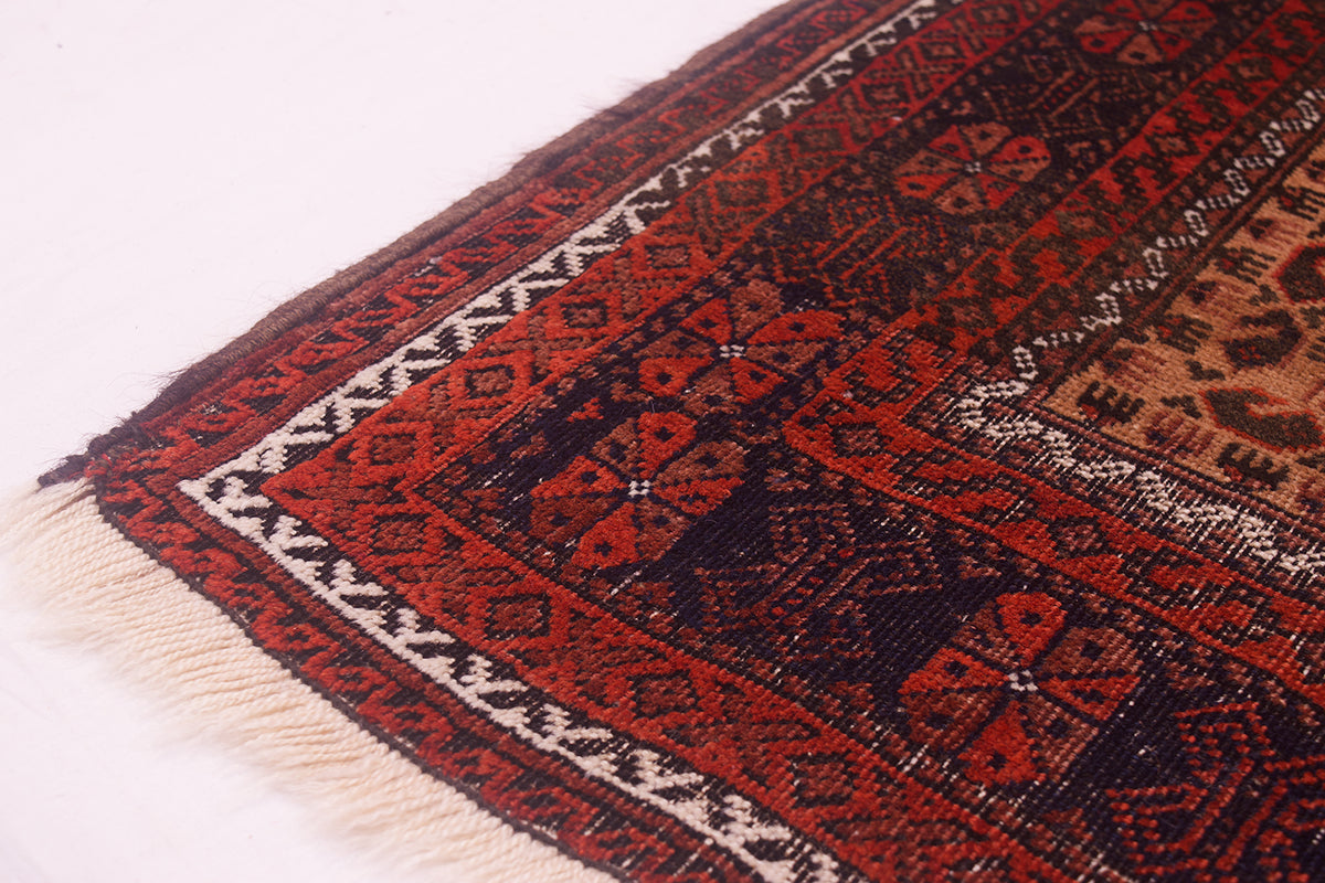 A close-up of a worn out patterned Belouch antique rug corner showing a dark red and black geometric design with cream fringe on a white background.