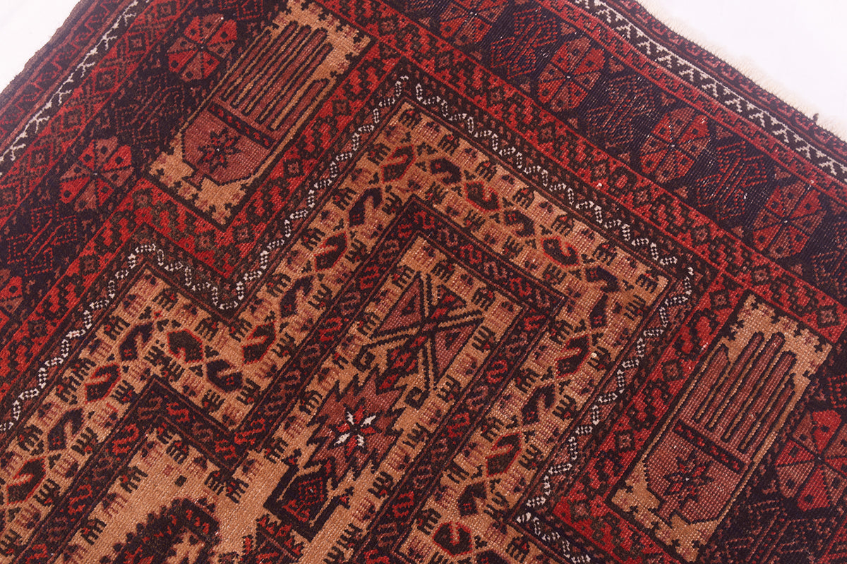 A close-up of a patterned antique Afghan Belouch prayer rug showing geometric and tribal-inspired motifs in deep red, terracotta, black, and beige.