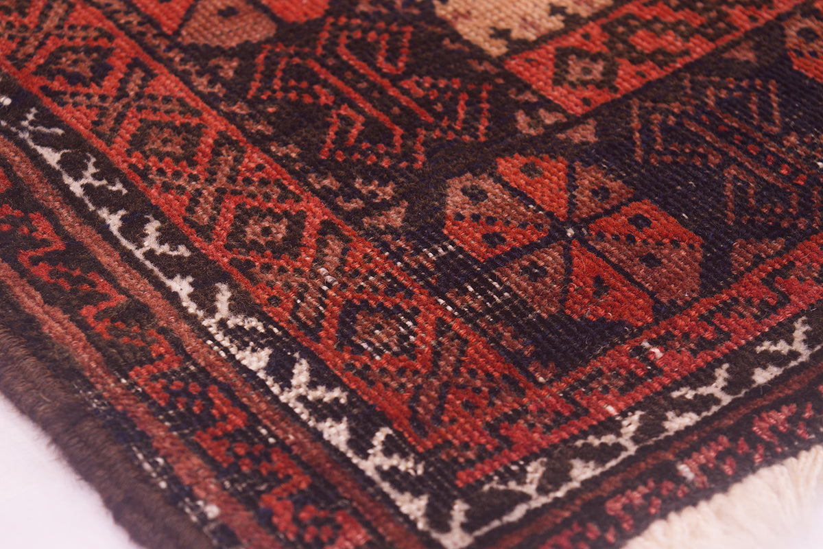 A close-up of the wear on a patterned Afghan Belouch antique rug with geometric tribal designs in red, black, and cream, showing the detailed woven texture and fringe on one side.