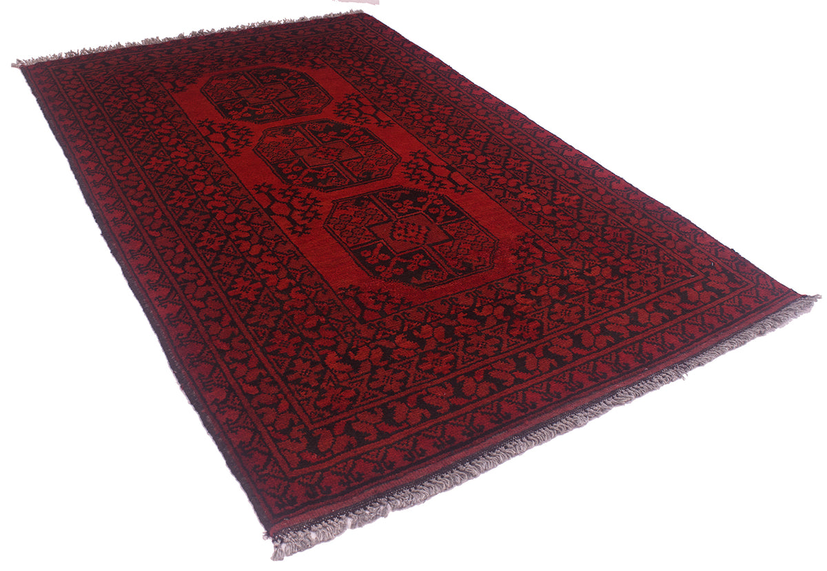 A rectangular red and black Afghan rug with fringed edges, shown at an angle on a plain white background.