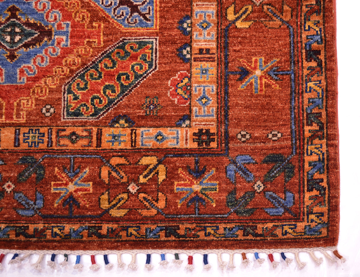 Close-up of a traditional rug corner showing a cinnamon red field with geometric motifs in blue, beige, yellow, and black, plus multicolour bound tassels along the edge.