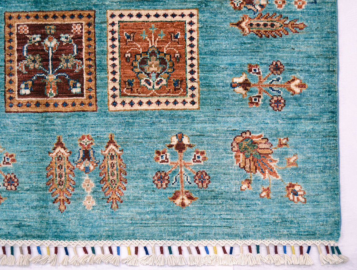 A close-up of a corner of a handmade Afghan Bakhtiari design turquoise carpet with traditional geometric and floral motifs in brown, beige, and cream, finished with a fringed edge.