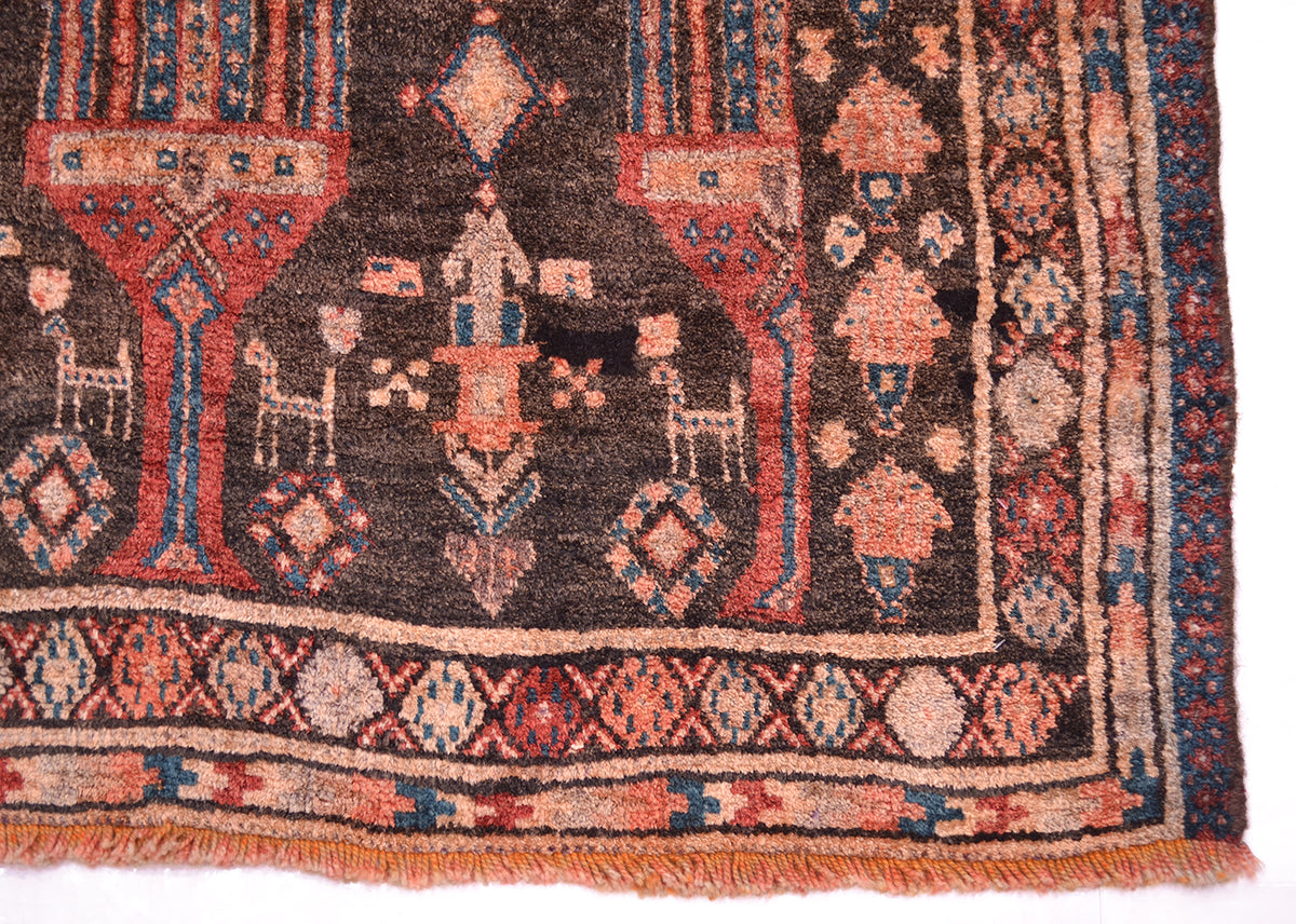 A close-up of an old patterned Belouch runner rug showing a dark brown field with red, beige, and blue geometric motifs and fringe along the bottom edge.