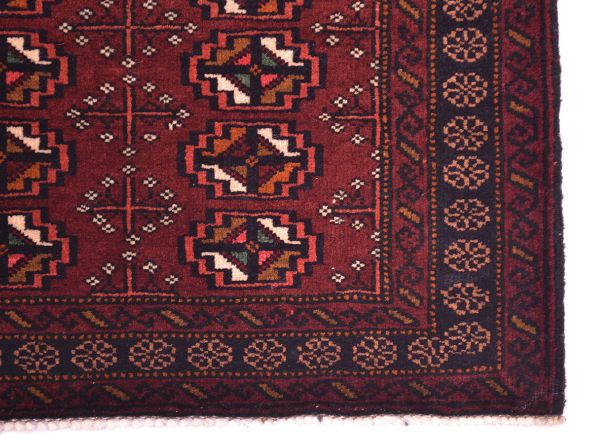 Close-up of a patterned small Afghan Belouch mat/rug corner showing a deep maroon field with repeating geometric medallions and a dark border with floral motifs.