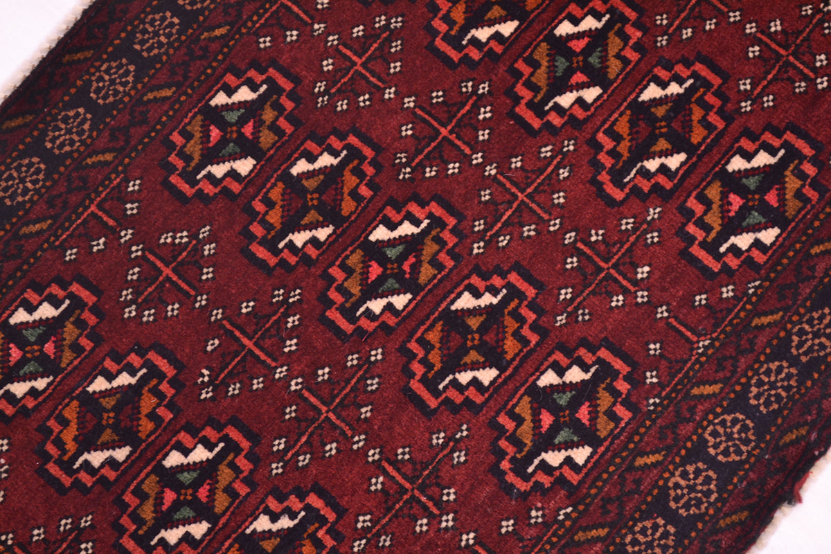 A close-up of a patterned Afghan Belouch small rug with a deep maroon field, featuring repeating geometric motifs in black, beige, orange, and green.