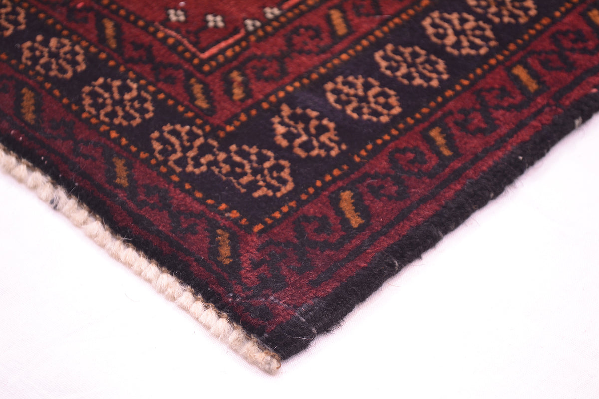 A close-up corner view of a patterned Maroon Afghan Belouch mat on a white background, showing a dark navy blue border with repeating beige floral motifs, a burgundy-red border, and a cream-coloured twisted fringe.