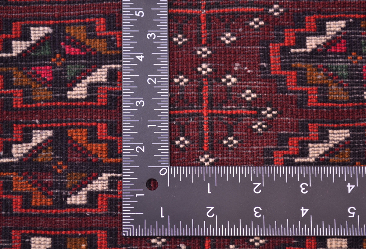 Close-up of the hand-knotted weave of a patterned Afghan Belouch mat with a dark maroon and black geometric design, shown alongside a metal ruler to show scale of the knotting.
