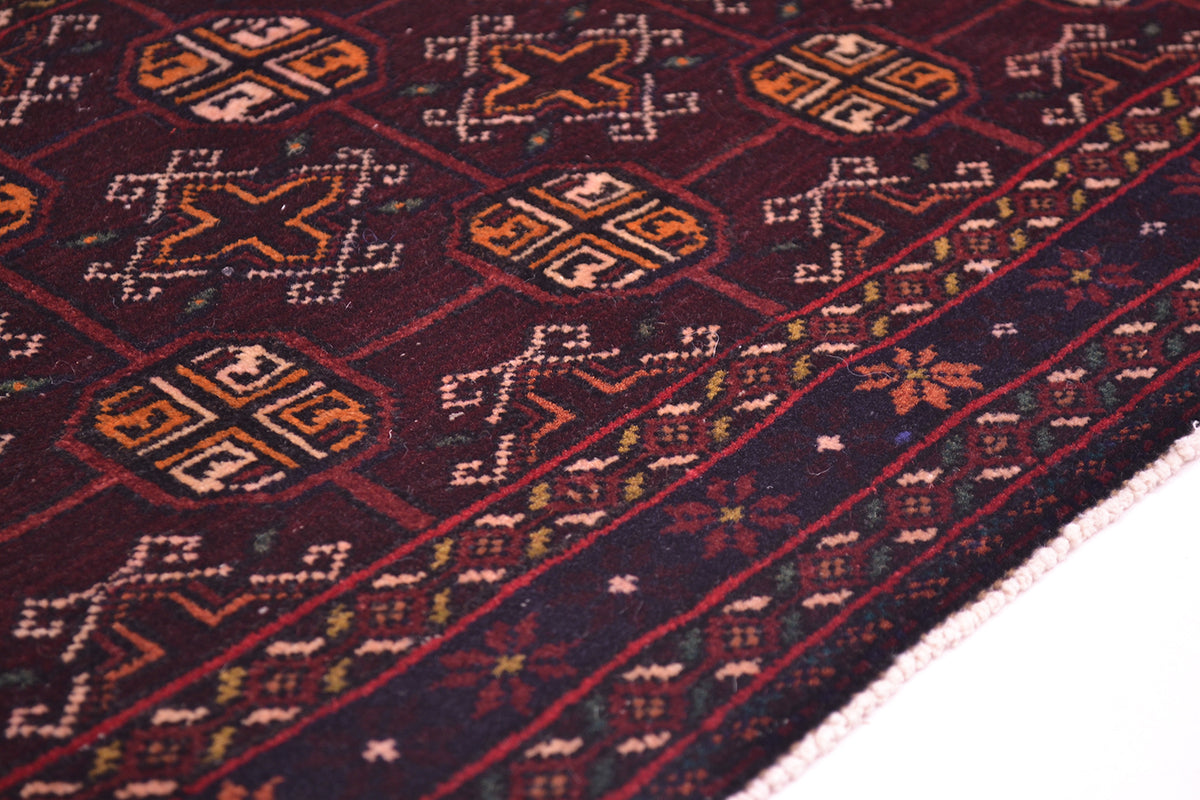 A close-up of a patterned Belouch Bokhara style mat with geometric motifs in deep burgundy, navy, orange, beige, and green.