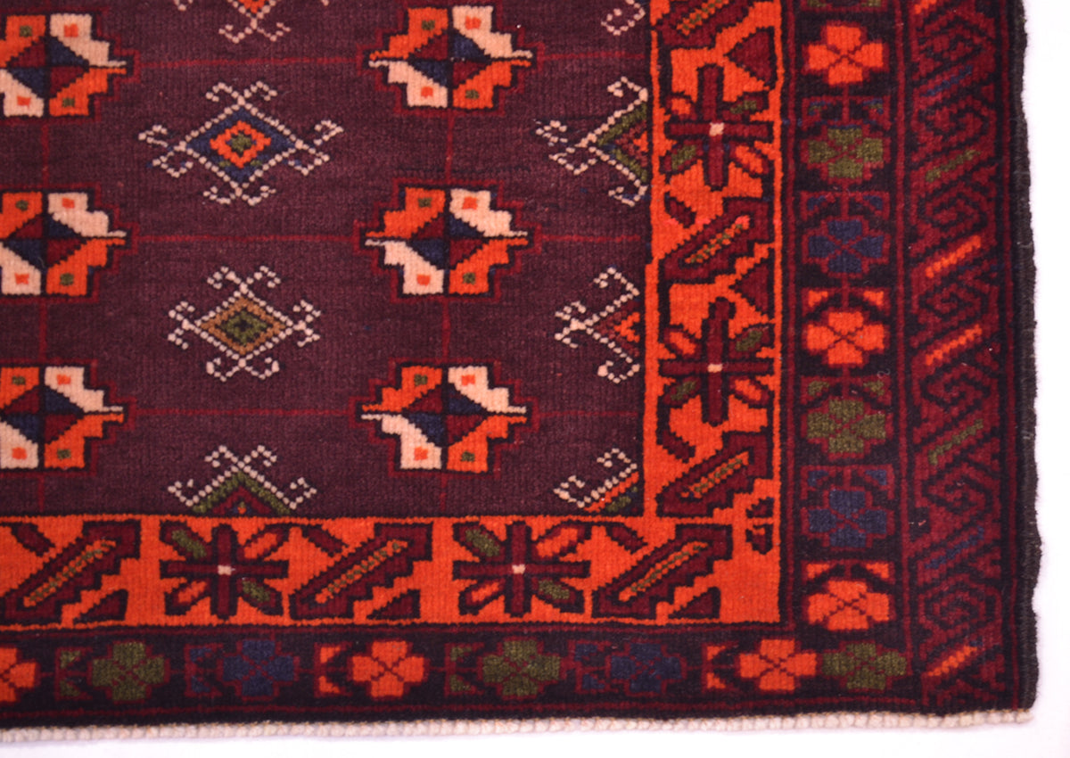 A close-up of a patterned rug corner showing a dark plum field with repeating geometric medallions in orange, cream, and green, plus a bold orange and dark border.