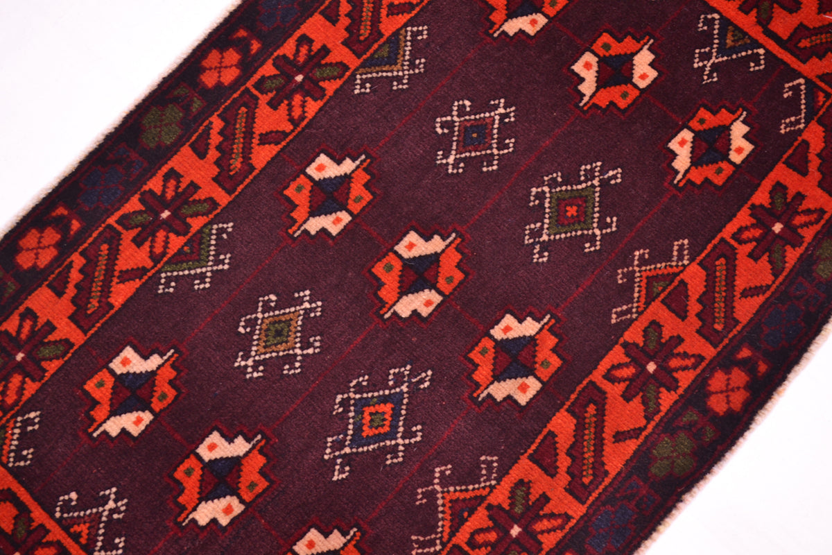 A close-up of a patterned rug showing a dark burgundy-brown field with repeating geometric motifs in red, orange, cream, and small olive green accents, bordered by red bands with decorative elements.