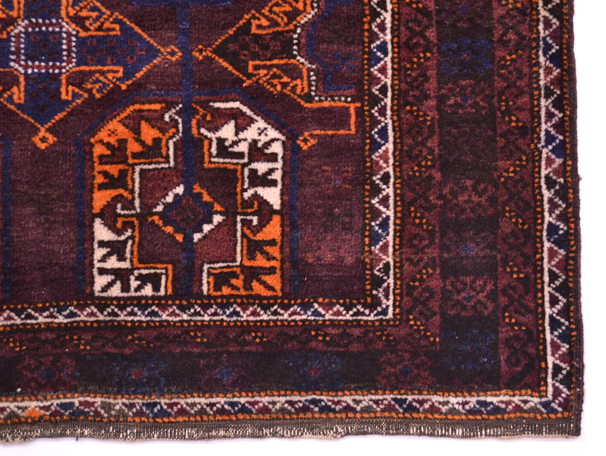 A close-up of an old tribal-style Belouch rug corner, showcasing intricate geometric and tribal patterns in deep burgundy, orange, navy blue, and cream.