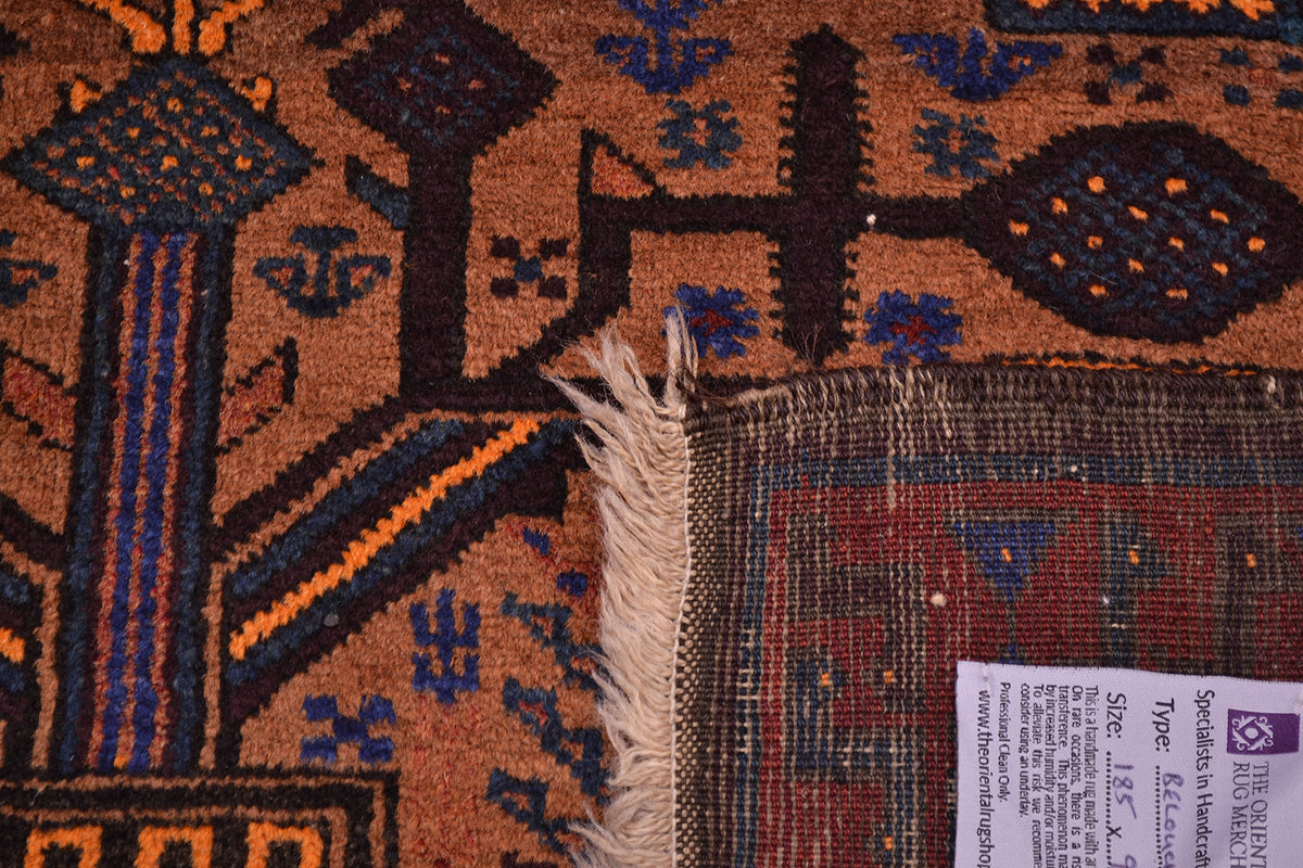 Close-up of a turned up corner of a patterned Afghan Belouch old rug showing a warm brown base with navy, orange, and beige geometric motifs, fringed edge, and part of a product tag.