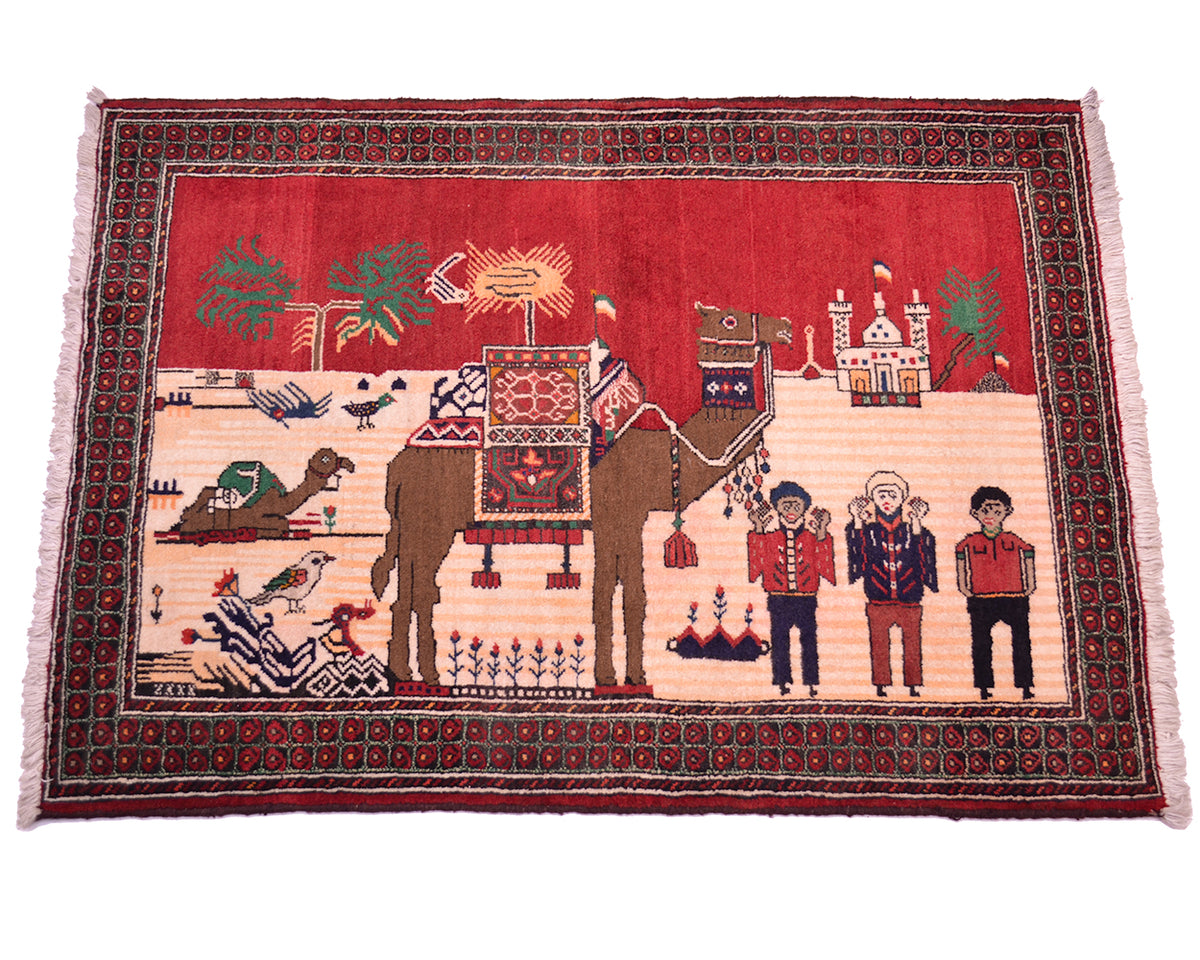 A rectangular handwoven Pictorial Belouch rug with a red and beige central scene featuring an camel, birds, and human figures, surrounded by a dark patterned border and pink fringe on a white background.