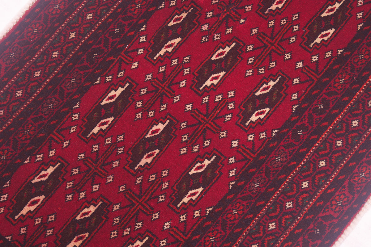 A close-up of a Afghan handmade red Belouch mat with repeating geometric gul motifs in dark brown, beige, and white.