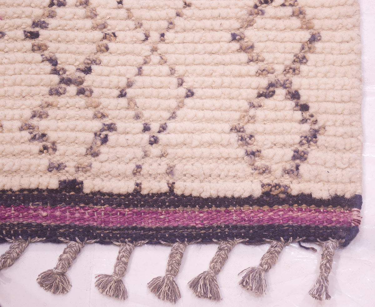 Close-up of a woven runner with a cream base, charcoal geometric patterning, a purple and dark stripe kilim border, and tasseled fringes along the bottom edge.