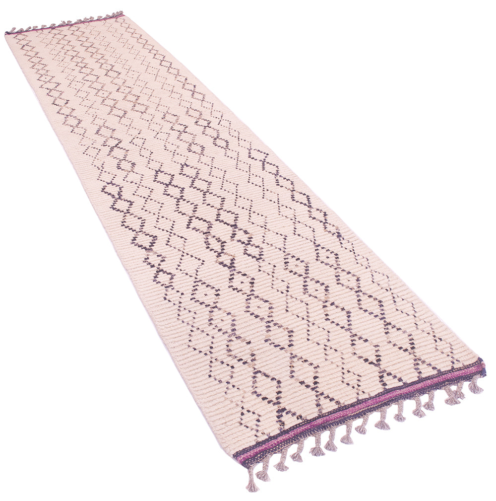 A long rectangular runner rug in a light cream tone with a repeating charcoal geometric diamond pattern and purple kilim ends with tassel fringe on both ends.