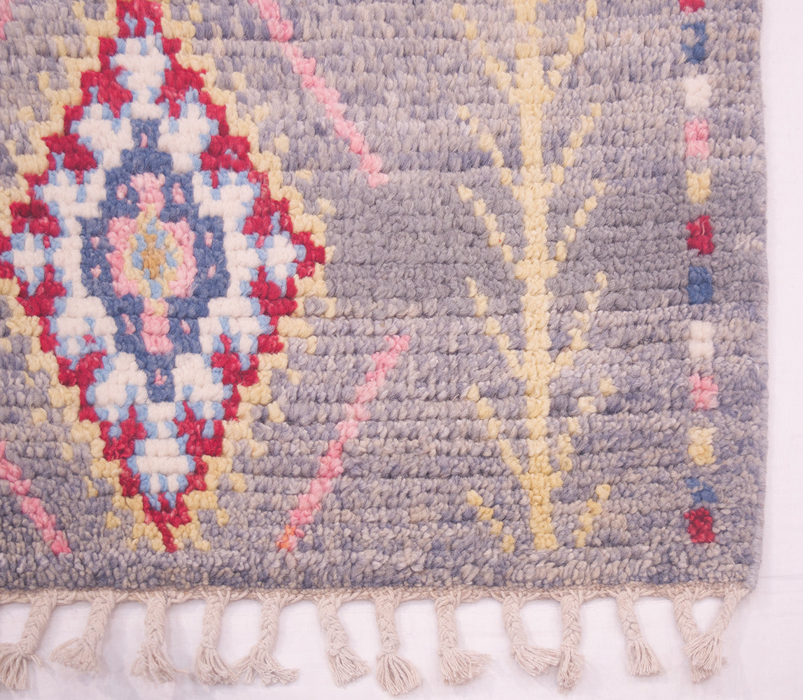 A close-up of a handwoven Moroccan-style Afghan rug corner showing a grey base with a multicolour diamond medallion pattern in red, cream, blue, and pink, plus a cream branch motif and fringe tassels along the bottom edge.