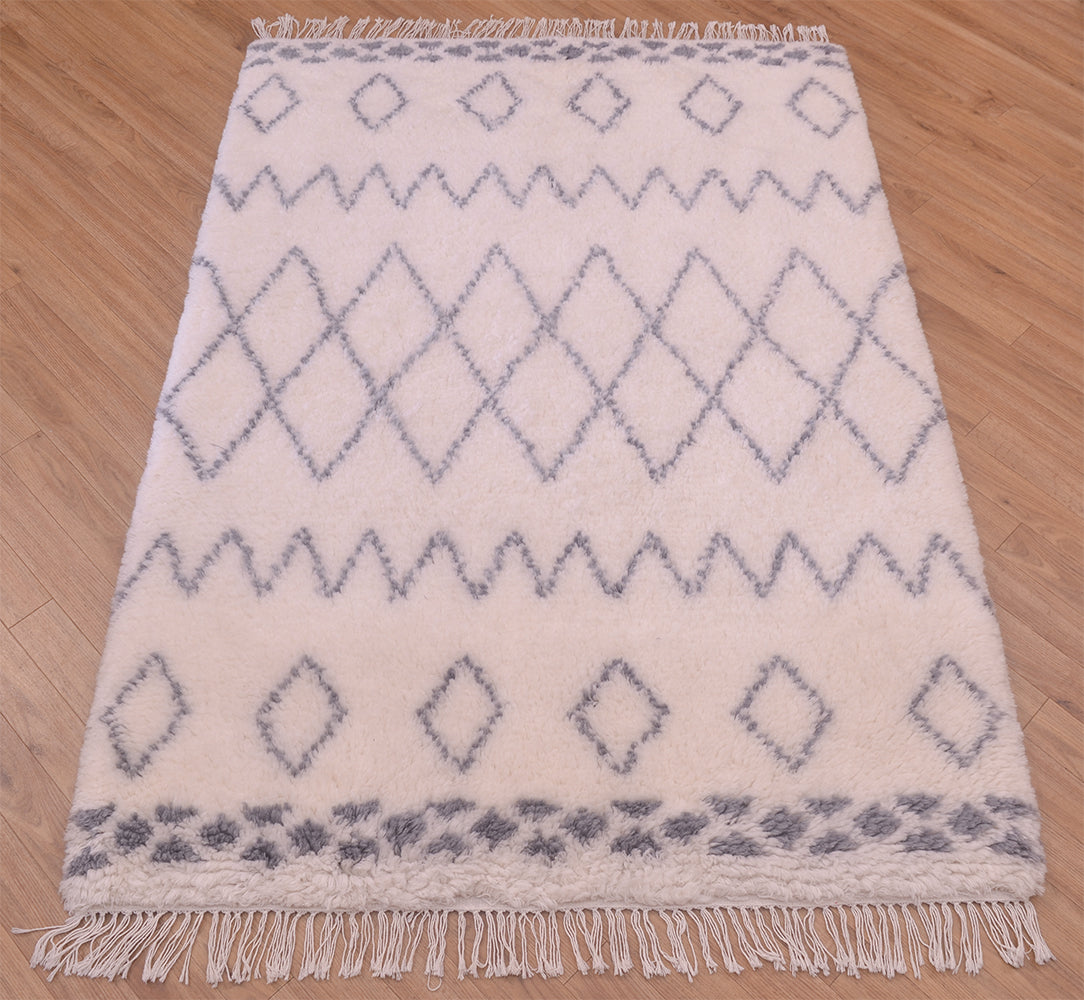 A rectangular woven shaggy pile Berber area rug with a cream base and grey geometric diamond and zigzag patterns, finished with fringe on the top and bottom edges, laid flat on a light wood floor.