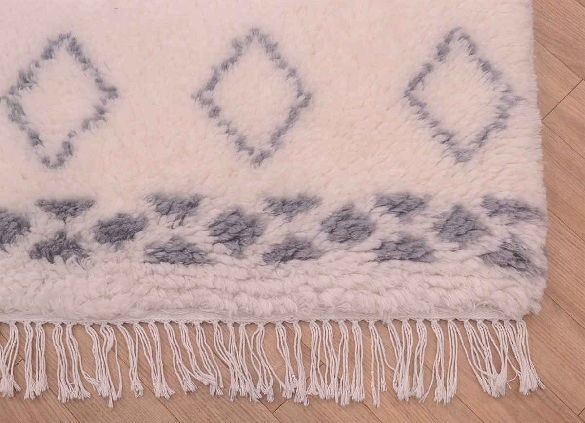 A close-up of a plush cream-colored shaggy pile Berber rug with a grey geometric diamond pattern and fringe tassels, laid on a light wood floor.
