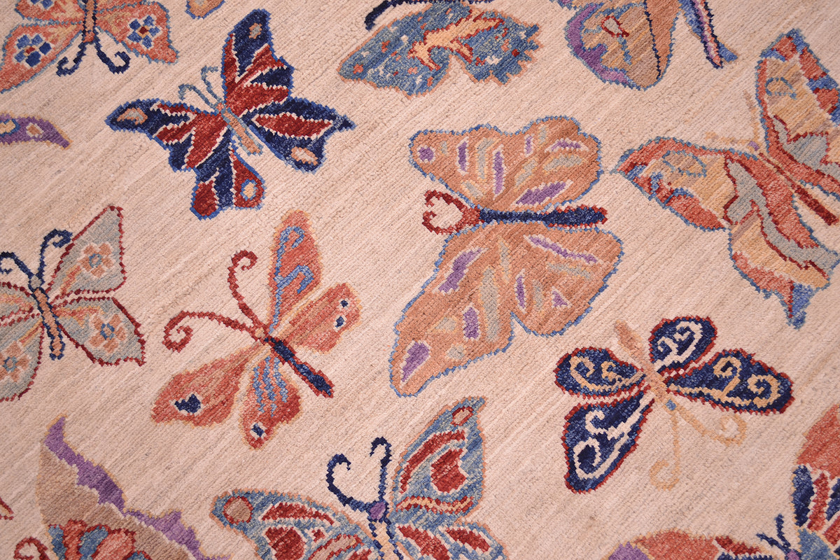 A close-up of a handwoven Afghan rug featuring a repeating pattern of stylised butterflies and moths in muted red, blue, purple, and peach tones on a warm beige background.
