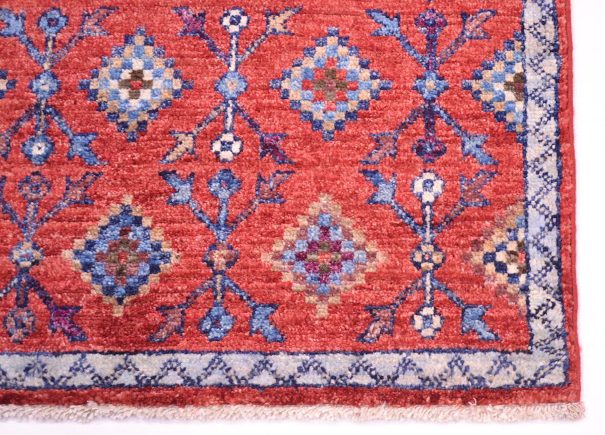 A close-up of a traditional patterned Chobi Afghan rug/mat corner, featuring a coral-red field with blue, beige, and purple geometric medallions and a cream border.