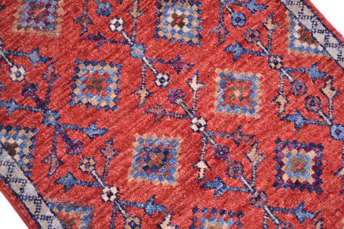 A close-up of a traditional tribal patterned small Afghan rug with a warm red-orange field, blue and beige geometric medallions, and floral motifs.