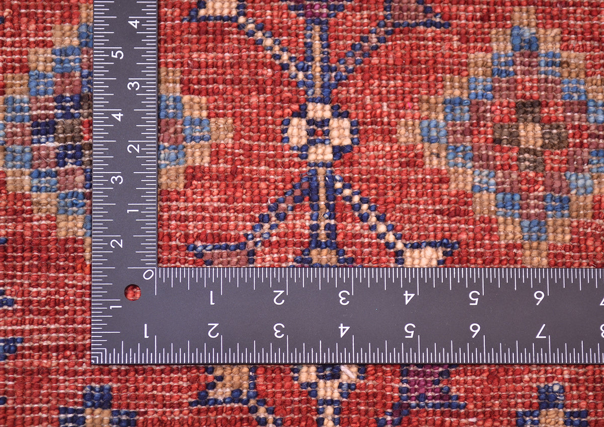 A close-up of the hand-knotted back of a patterned Afghan Chobi tribal design mat in red, blue, beige, and brown tones, shown with a metal ruler to indicate scale of the knotting.