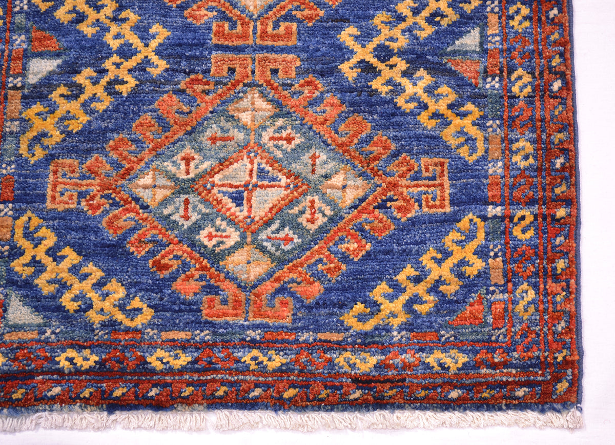A close-up of a patterned Afghan short runner rug corner showing a blue field with orange, red, and cream geometric motifs and fringe along the bottom edge.