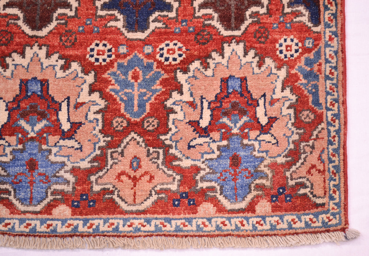 A close-up of a corner of a traditional patterned Persian style Chobi rug showing a red field with blue, beige, and brown floral motifs and a fringed edge.