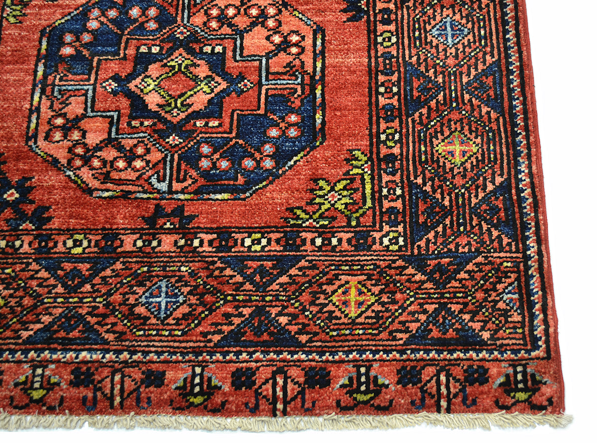 A close-up of a traditional handwoven Afghan Ersari runner corner showing its intricate geometric patterns, coral-red field, navy blue borders, and cream fringe.