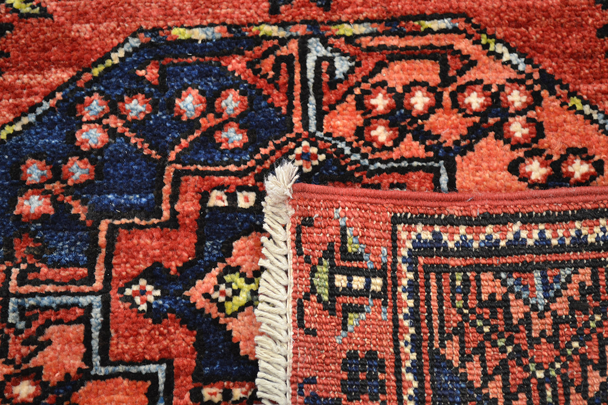 Close-up of the hand-knotted back of a patterned Afgha runner with the corner turned over with a rust red base, dark blue geometric sections, and cream fringe at the corner.