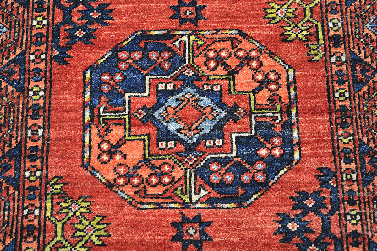 A close-up of a traditional handwoven Afghan runner rug featuring a vibrant red field with a central octagonal medallion in navy blue, orange, and light blue, surrounded by floral motifs and patterned borders.