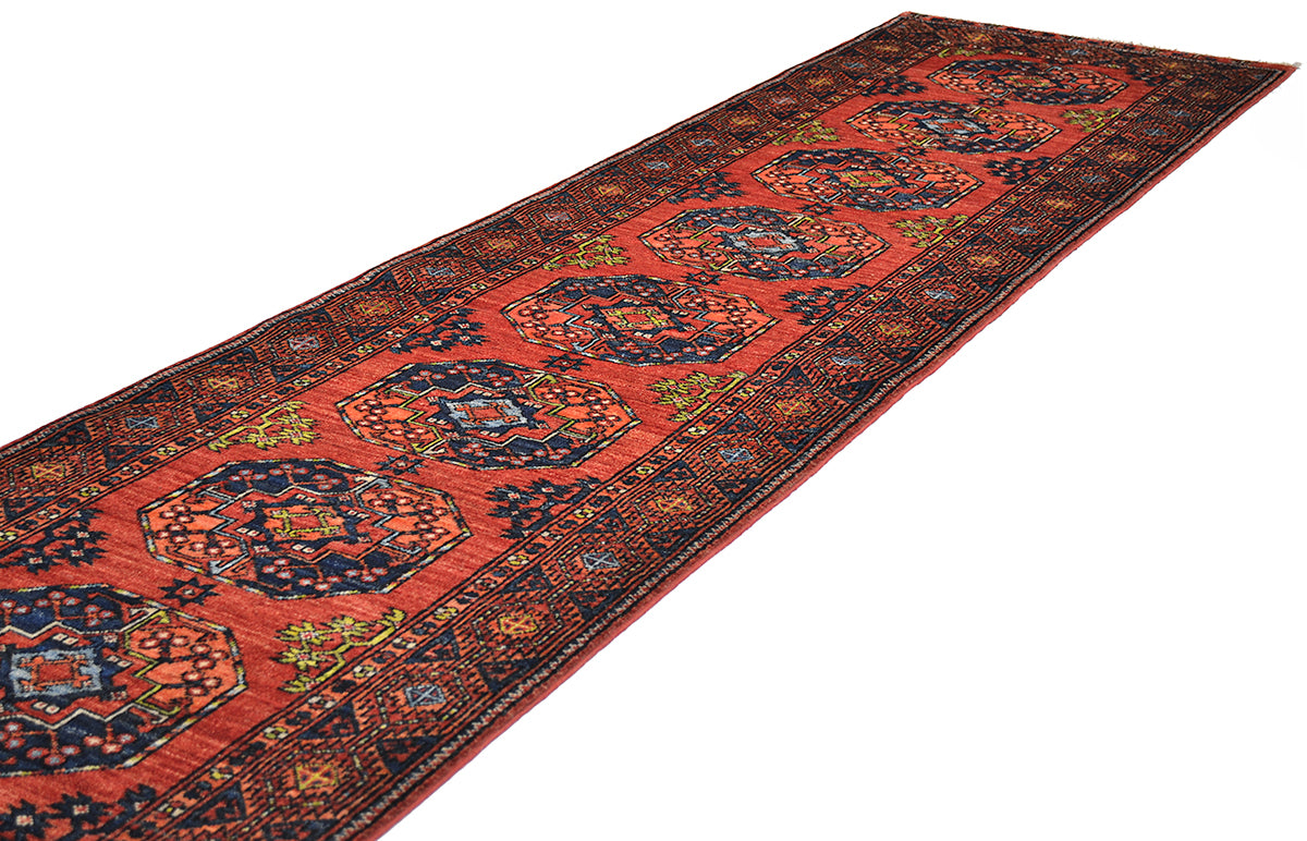 A long oriental Afghan corridor runner rug with a warm rust field, dark navy blue central medallions, and intricate multicolour floral patterns, shown diagonally on a white background.