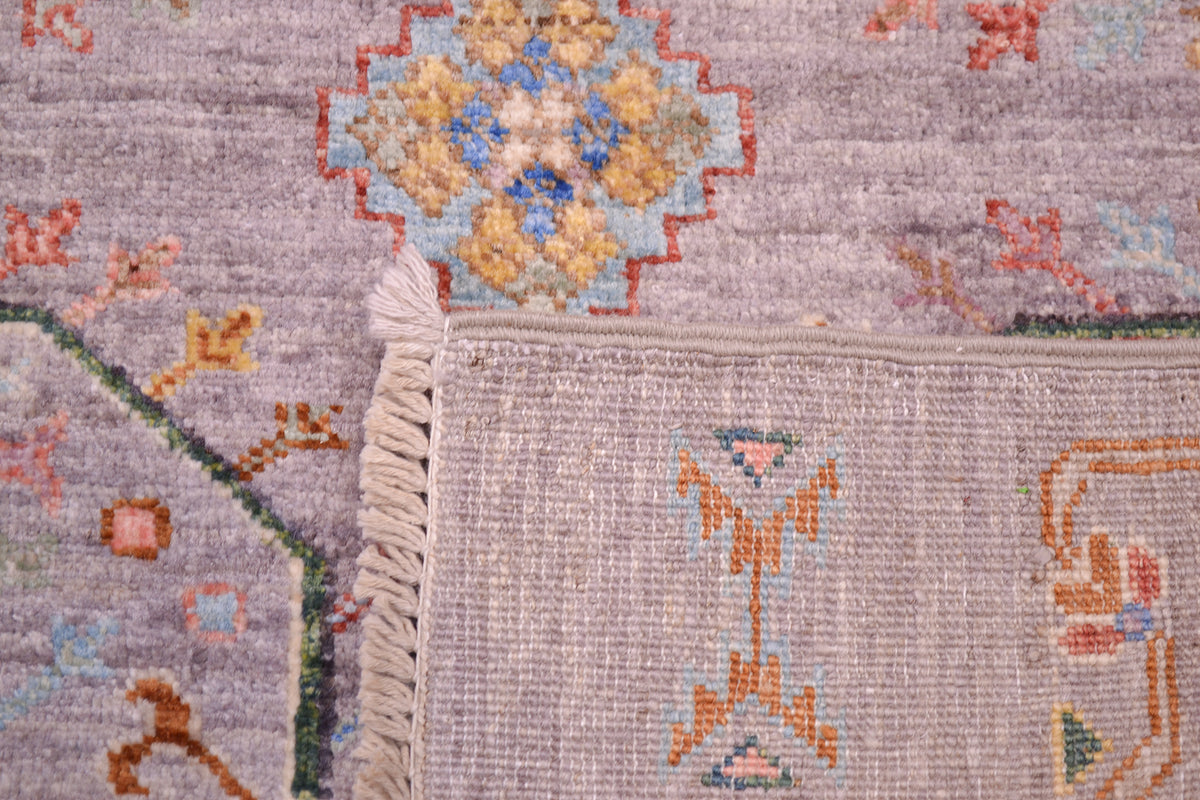 Close-up of a woven Afghan rug corner showing cream fringe, a grey base, and multicoloured floral and geometric motifs in beige, orange, blue, and green.