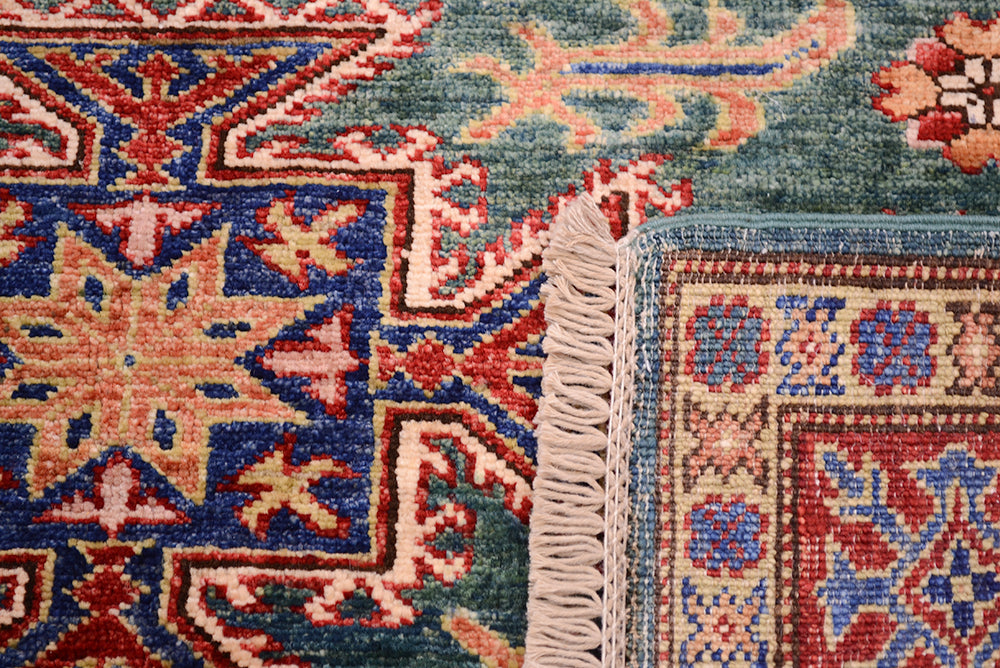 Close-up of a traditional patterned rug showing corner details, including a cream fringe edge, with a teal-blue field, red and beige motifs, and blue border sections.