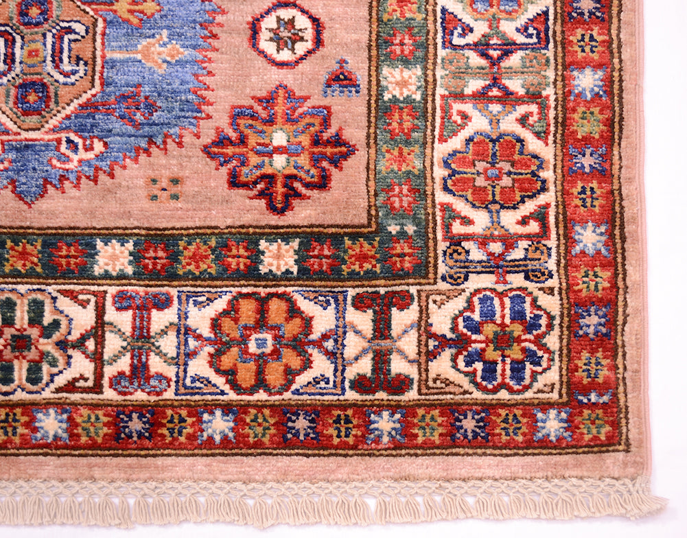 A close-up of a traditional patterned Afghan Kazak rug corner showing a beige field, red and blue geometric motifs, decorative borders, and cream fringe.