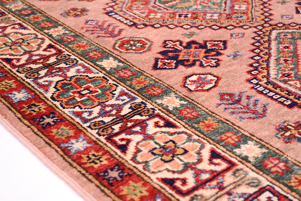 A close-up of a traditional Kazak patterned rug with a dusty pink field and detailed borders in red, blue, green, and cream.